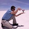 A man kneels on a vast, white salt flat during his incredible adventure, holding his hands to make a pink flamingo in the distance appear small enough to fit between his fingers, creating an optical illusion.