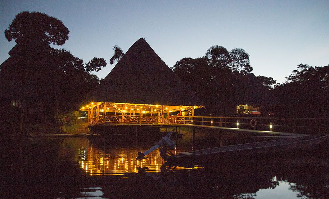 A thatched-roof, open-air building at Napo Wildlife Center Eco-Lodge glows with warm lights at dusk, its reflection shimmering on calm water in the Ecuador Amazon. A small boat is docked in the foreground, surrounded by trees.
