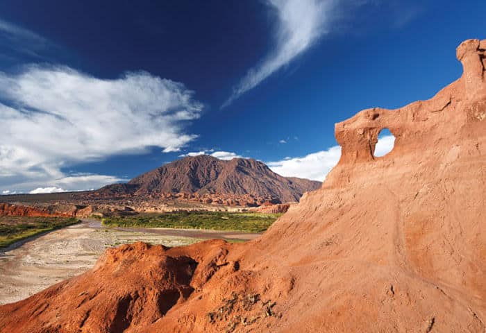 Red rock formations with a natural arch in the foreground, backed by a mountain, green vegetation, and a partially cloudy blue sky—one of the stunning Argentina attractions perfect for any Argentina travel adventure.