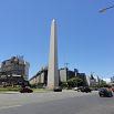 A tall stone obelisk stands in the center of a wide city intersection, surrounded by cars and modern buildings under a clear blue sky—a striking landmark that promises a memorable adventure for those who visit.