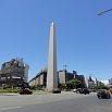 The image shows a tall, narrow obelisk standing in the center of a busy city intersection, surrounded by cars and buildings under a clear blue sky—a perfect spot to experience during a fantastic trip.