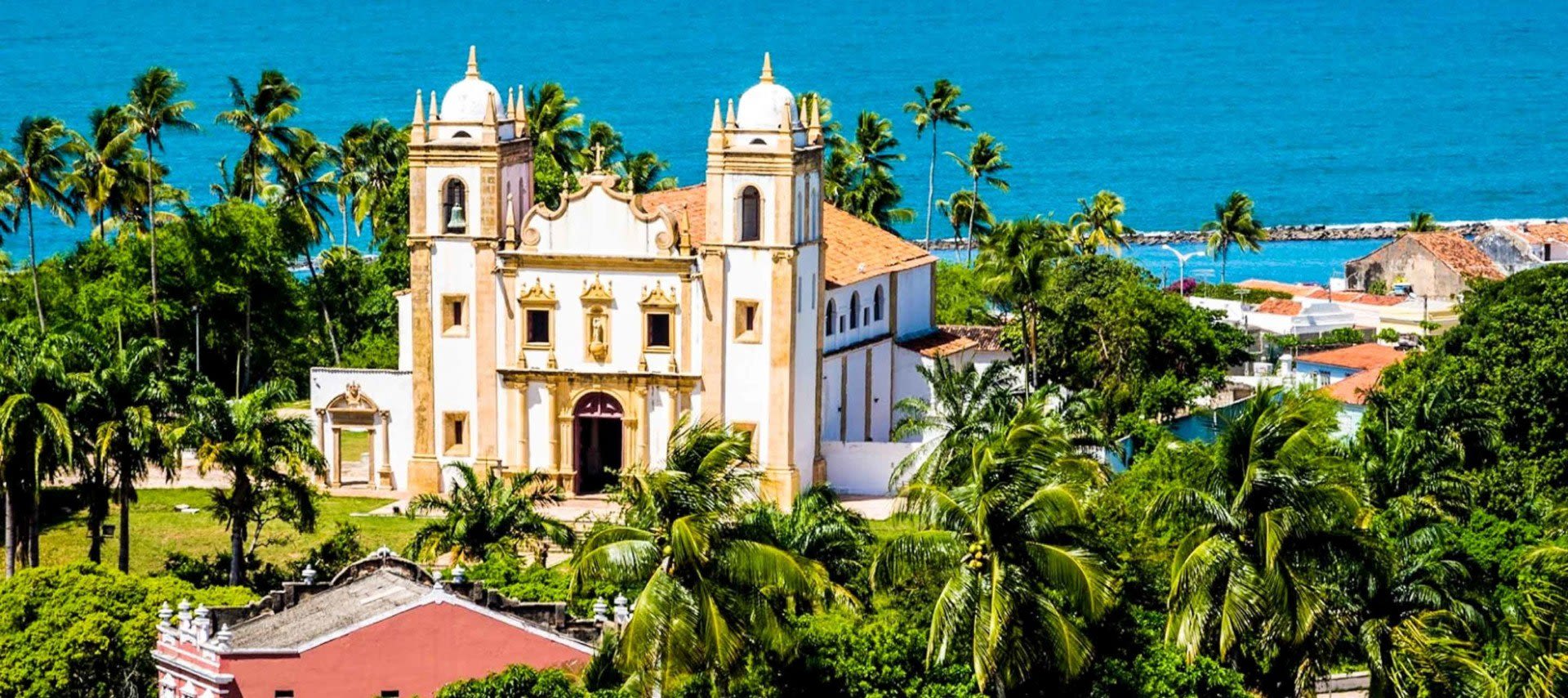 A historic colonial church with two bell towers stands among lush palm trees near the blue ocean under a clear sky, with other colorful buildings nearby—a picture-perfect stop on Olinda Tours.