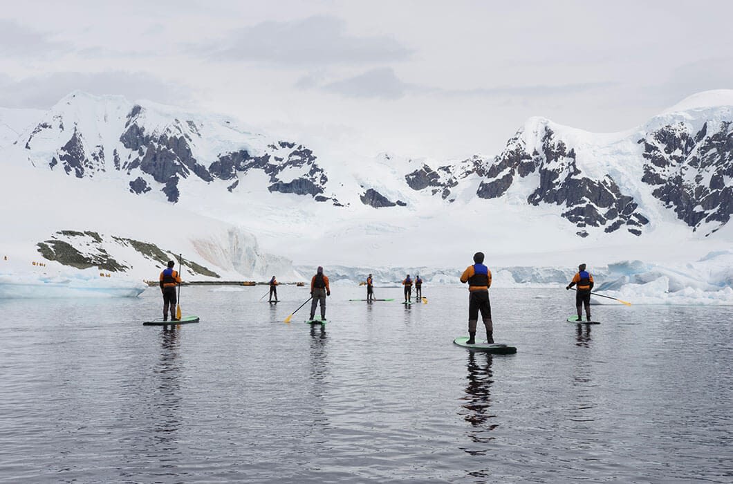 A group of people paddleboarding on calm water in a snowy, mountainous landscape—one of the unique Antarctica activities—surrounded by ice and snow-covered peaks under a cloudy sky.