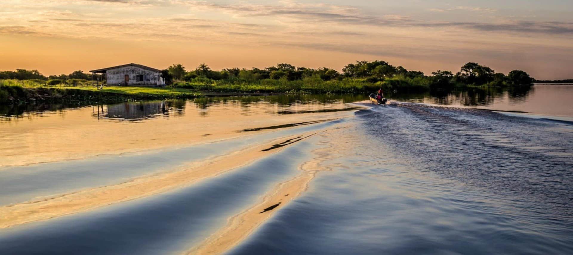 A small boat creates ripples on a calm river at sunset, reflecting golden and blue hues. Lush greenery and a rustic building line the riverbank—an inviting scene for a true Paraguay Adventure under a partly cloudy sky.