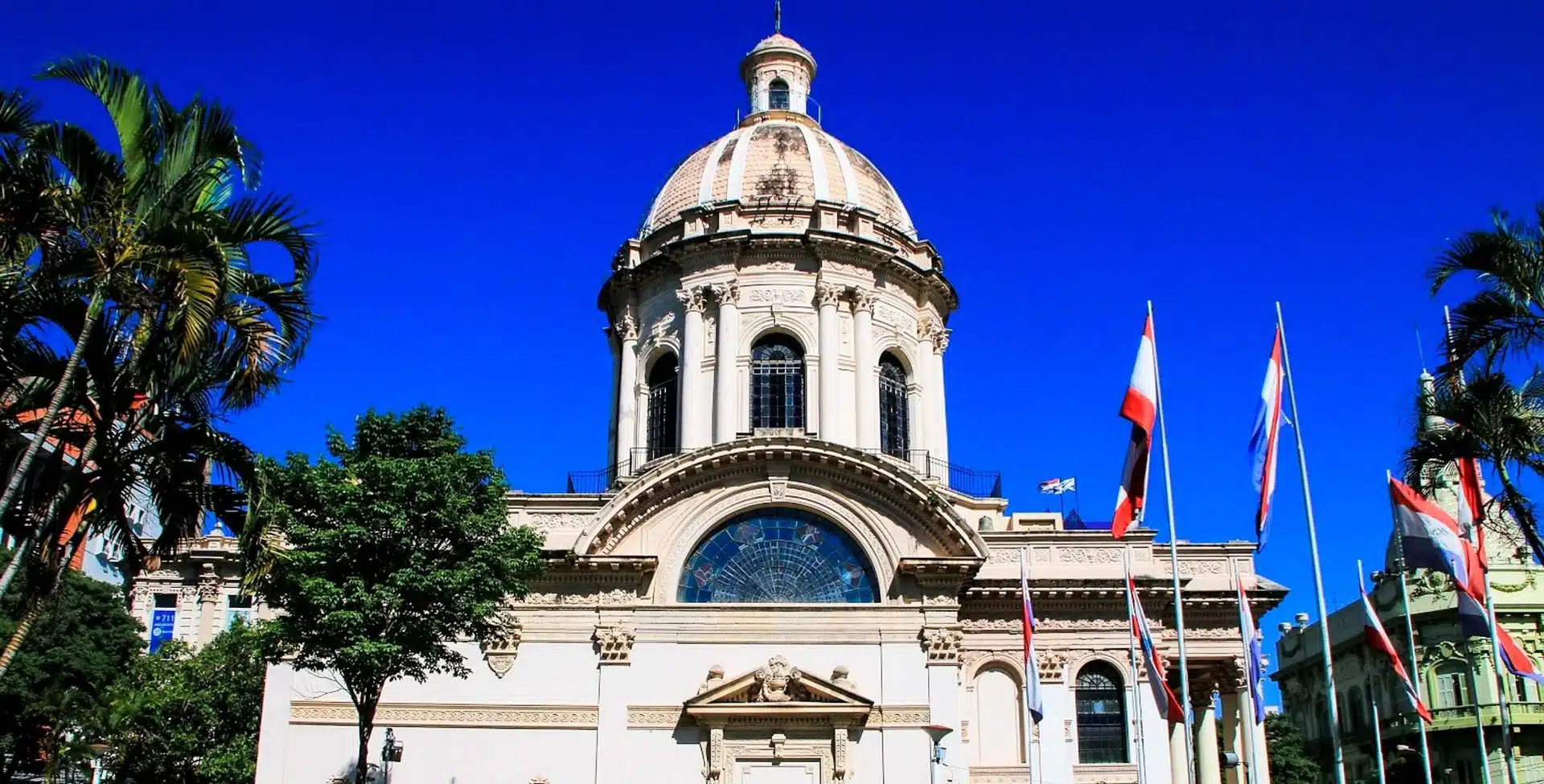 A neoclassical building with a large central dome, arched windows, and several national flags in front stands proudly beneath a bright blue sky and palm trees—a must-see Paraguay landmark for any Travel Guide.