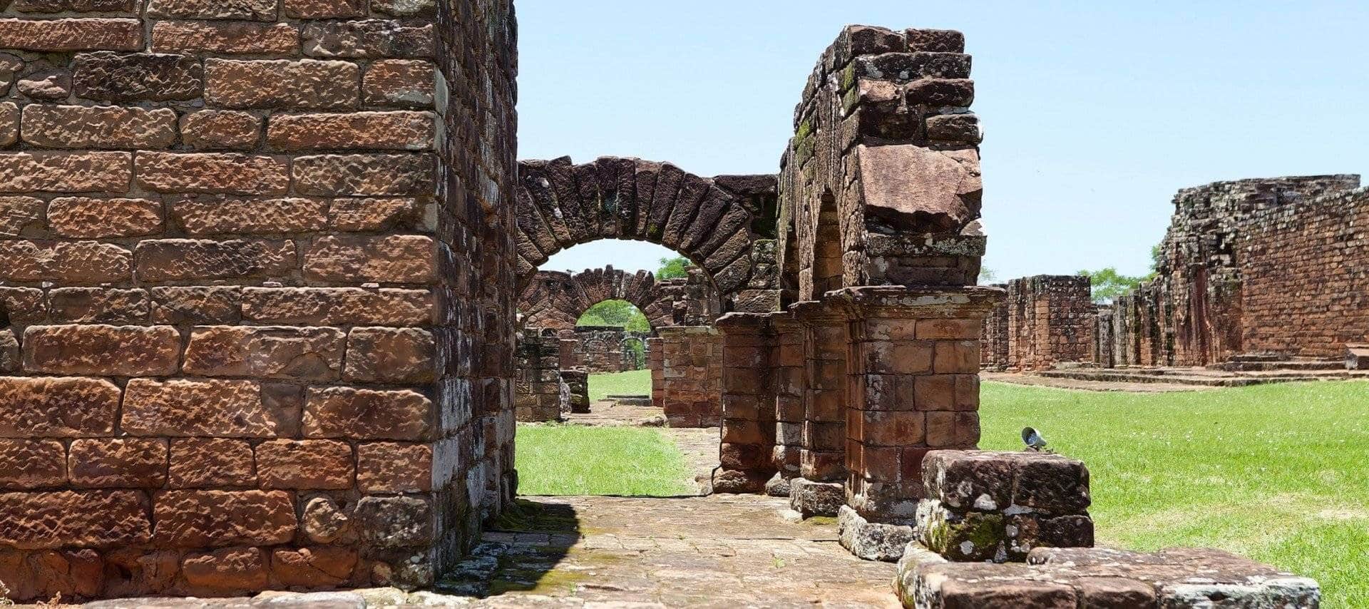 Ancient stone ruins with arches and weathered walls stand on a grassy field under a clear blue sky, inviting visitors to explore this UNESCO site in Paraguay and discover its rich history.