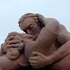 A close-up of a stone sculpture showing two intertwined human figures, one holding the other's head and back, both displaying intense facial expressions against a plain sky—a powerful scene you might encounter on a South America travel adventure.