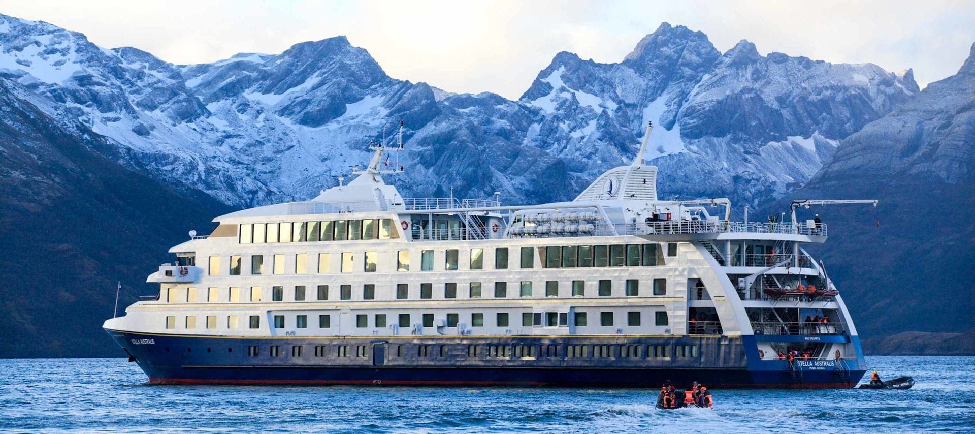 A large white and blue cruise ship sails on calm water with snow-covered mountains in the background. Small inflatable boats with passengers explore nearby—a quintessential Patagonia Cruises adventure.