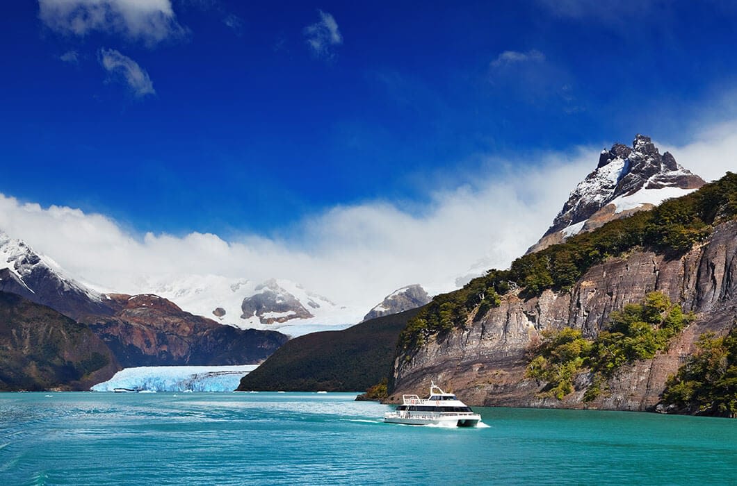 A white boat sails on turquoise water surrounded by rocky, forested cliffs and snow-capped mountains, with a glacier in the background—an unforgettable scene among top Chile attractions under a blue sky with scattered clouds.
