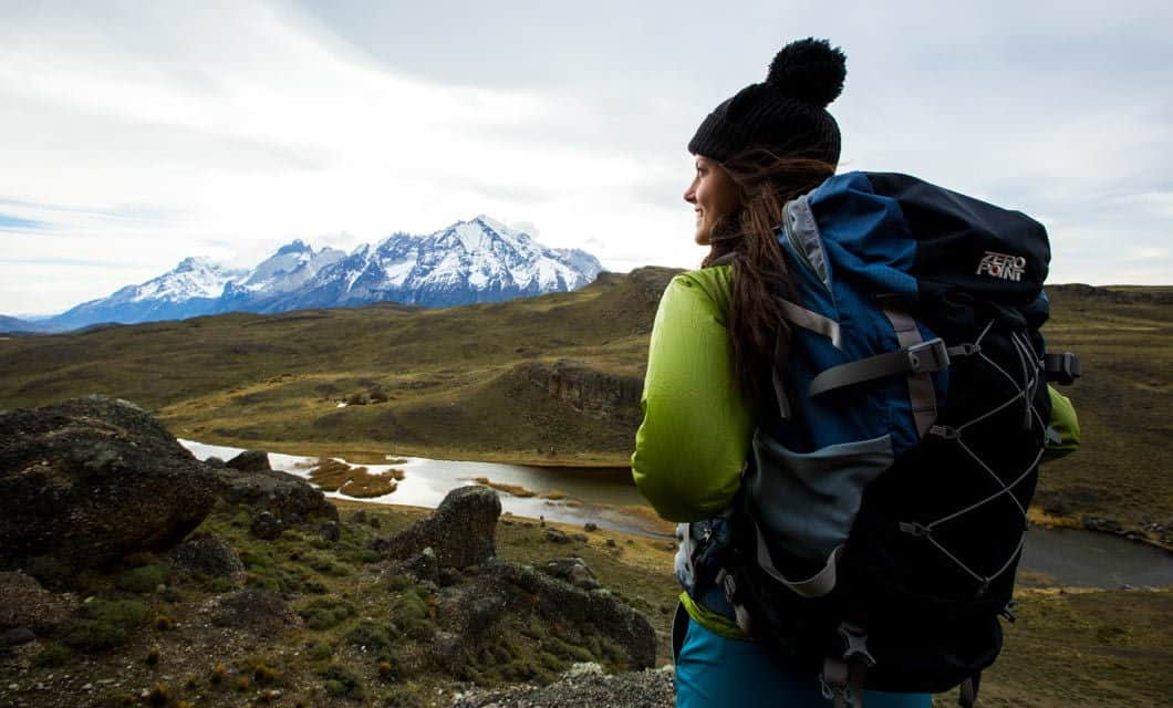 A person wearing a green jacket and black beanie with a large backpack stands on a rocky hillside, gazing at snow-capped mountains and a winding river near the renowned Tierra Patagonia hotel under a cloudy sky.