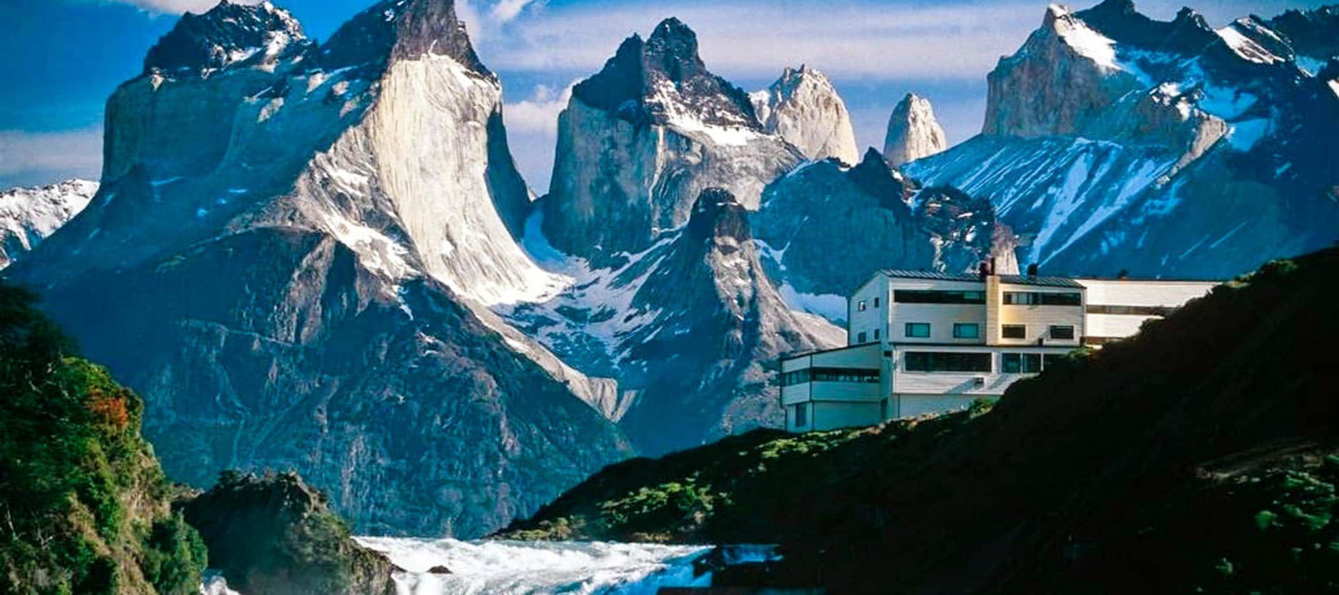 A modern white building sits on a hill, resembling the famed Patagonia lodges, with dramatic jagged mountains and snow patches rising in the background under a bright blue sky.