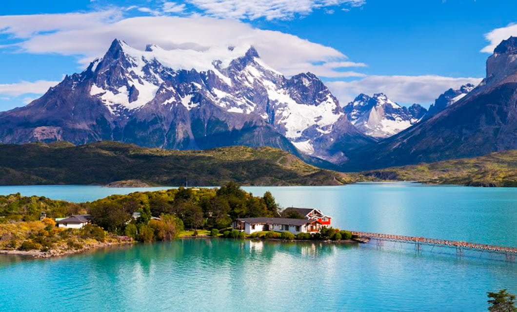 A picturesque luxury lodge sits by a turquoise lake, with a wooden bridge connecting it to land. Snow-capped mountains and lush green hills rise in the background under a bright blue sky with scattered clouds.