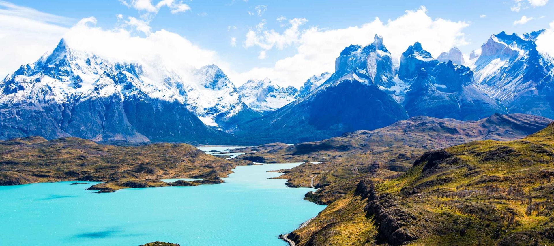 A turquoise lake winds through a rugged landscape with green hills and dramatic, snow-capped mountain peaks—an iconic scene from Torres del Paine, perfect for unforgettable Patagonia tours under a bright blue sky with scattered clouds.