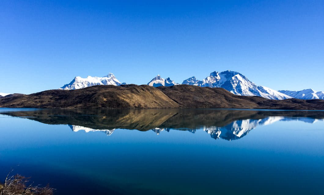 Snow-capped mountains and brown hills are reflected in a calm, blue lake beneath a clear sky, just steps from the luxurious Tierra Patagonia hotel and its renowned Patagonia spa.