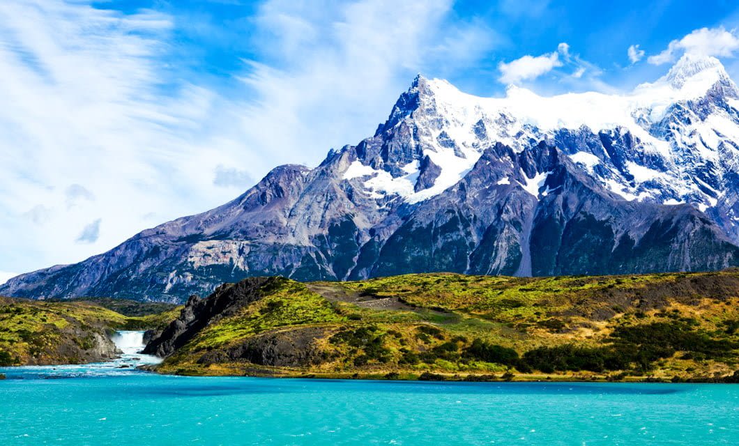 Snow-capped mountains rise above green hills and a turquoise lake in Patagonia under a bright blue sky with wispy clouds; a waterfall flows from the hills into the lake near an inviting Eco-Camp.