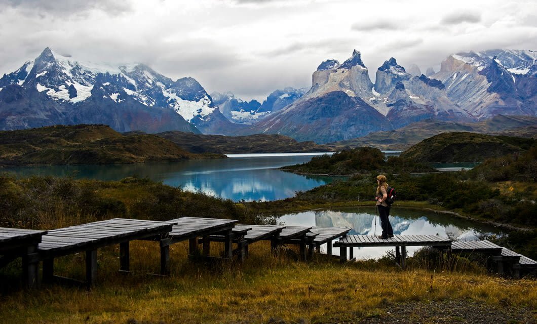A person stands on a wooden walkway by a reflective lake, surrounded by grass and hills, with dramatic snow-capped mountains and a cloudy sky near the renowned Singular Patagonia Hotel.