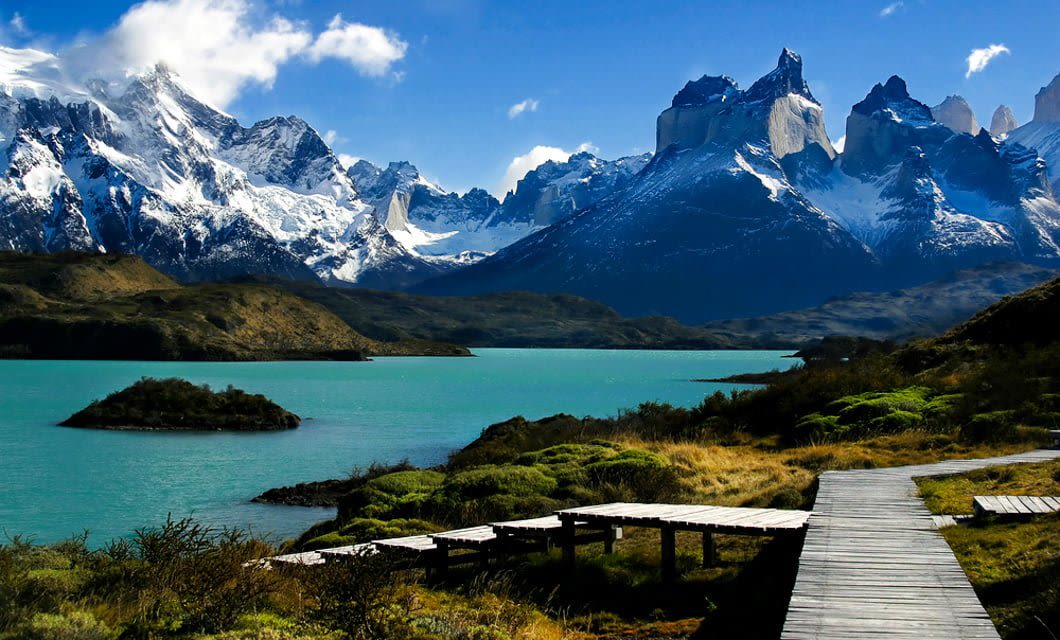 A wooden pathway leads toward a turquoise lake, surrounded by green vegetation, with dramatic snow-capped mountains in the background—just steps from the luxury Singular Patagonia Hotel under a partly cloudy blue sky.