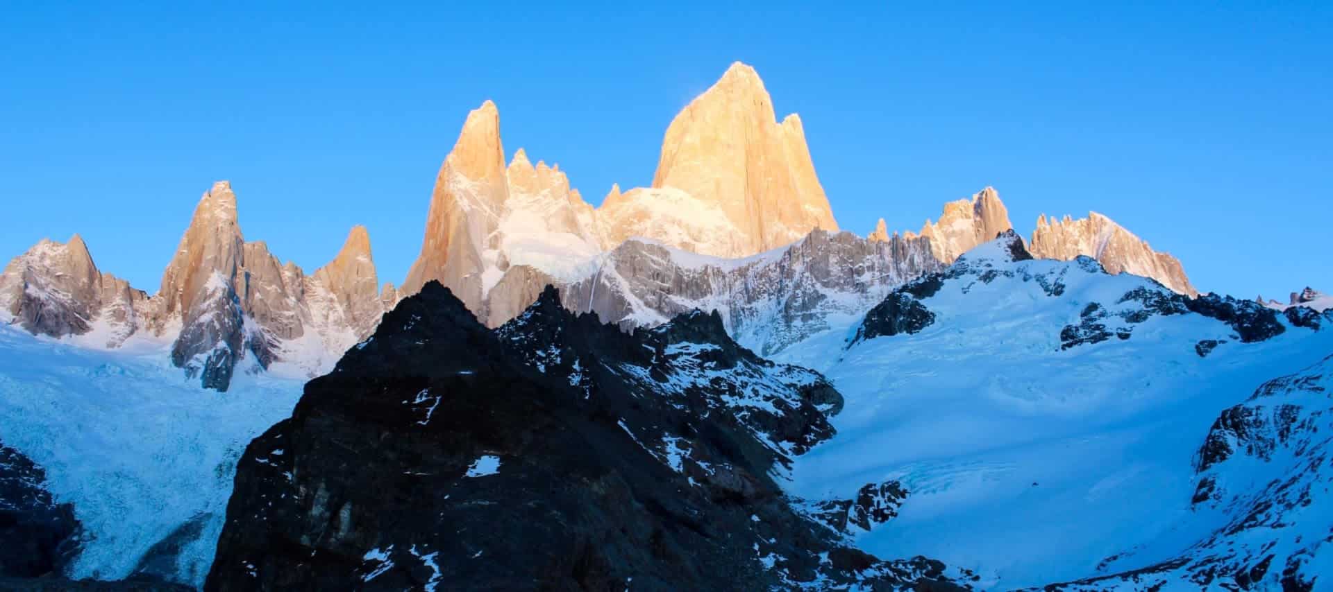 Jagged, snow-covered mountain peaks rise sharply under a clear blue sky. The tallest peak is bathed in golden sunlight, offering a breathtaking view perfect for Patagonia Tours and unforgettable adventure tours in this stunning region.