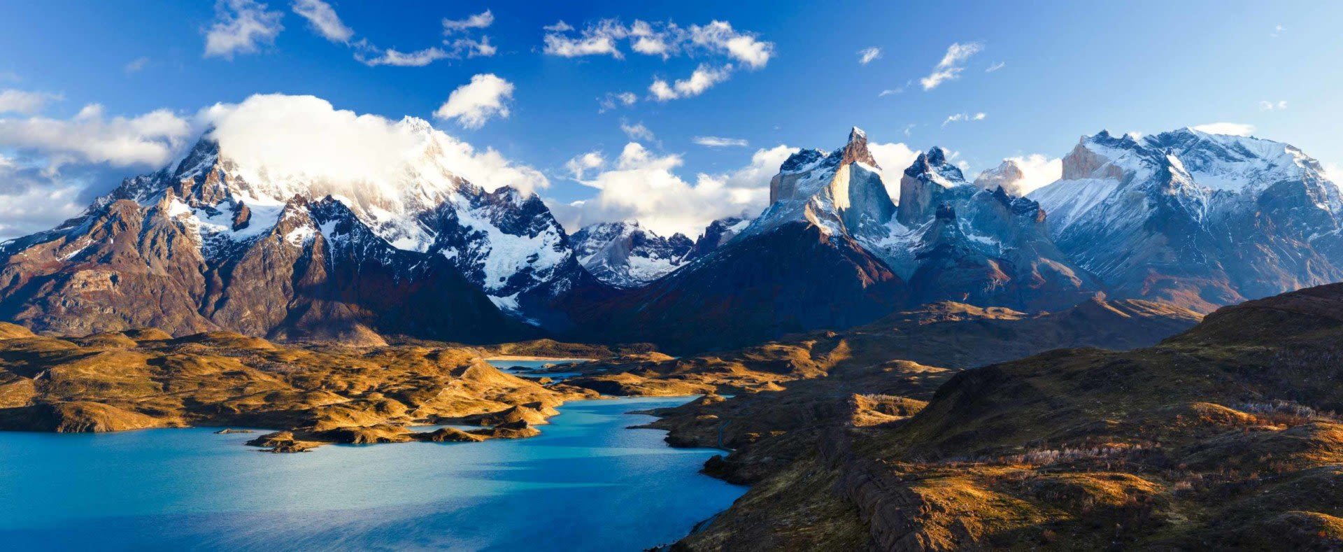 Snow-capped mountains rise above rugged hills and bright blue lakes under a clear sky in Patagonia, with scattered clouds clinging to mountain peaks in a dramatic, sunlit landscape.