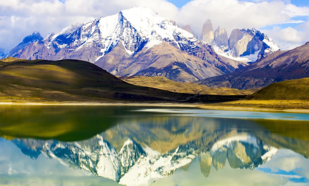Snow-capped mountains reflected in a calm lake, with green hills and blue sky with scattered clouds in the background—an idyllic scene near Patagonia Camp, capturing the wild beauty of Patagonia.