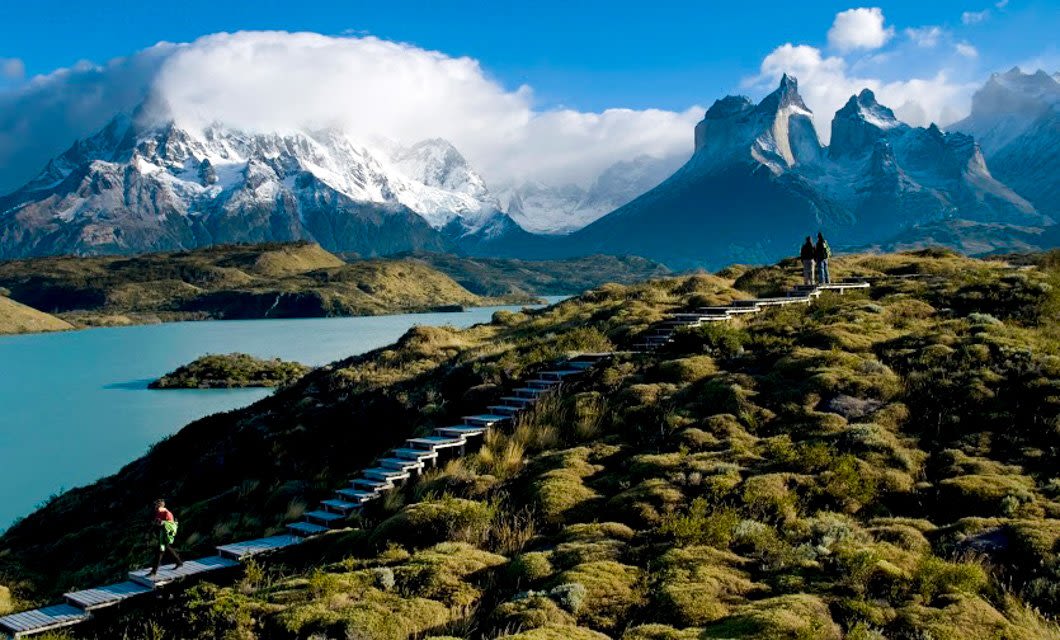 A wooden walkway winds through green hills toward turquoise water and jagged, snow-capped mountains under a partly cloudy blue sky—an iconic scene of adventure travel in Patagonia, with people walking along the path.