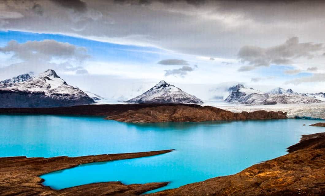 Turquoise blue lake bordered by rocky land, with snow-capped mountains and glaciers in the background under a cloudy sky—an iconic scene from Estancia Cristina in Patagonia, Argentina.