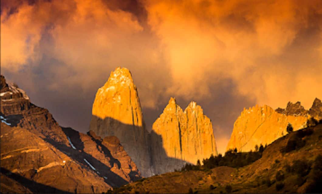 Dramatic orange clouds fill the sky above sharp, rocky mountain peaks, partially lit by golden sunlight, with dark ridges and sparse vegetation in the foreground near an Eco-Camp Patagonia Lodge.