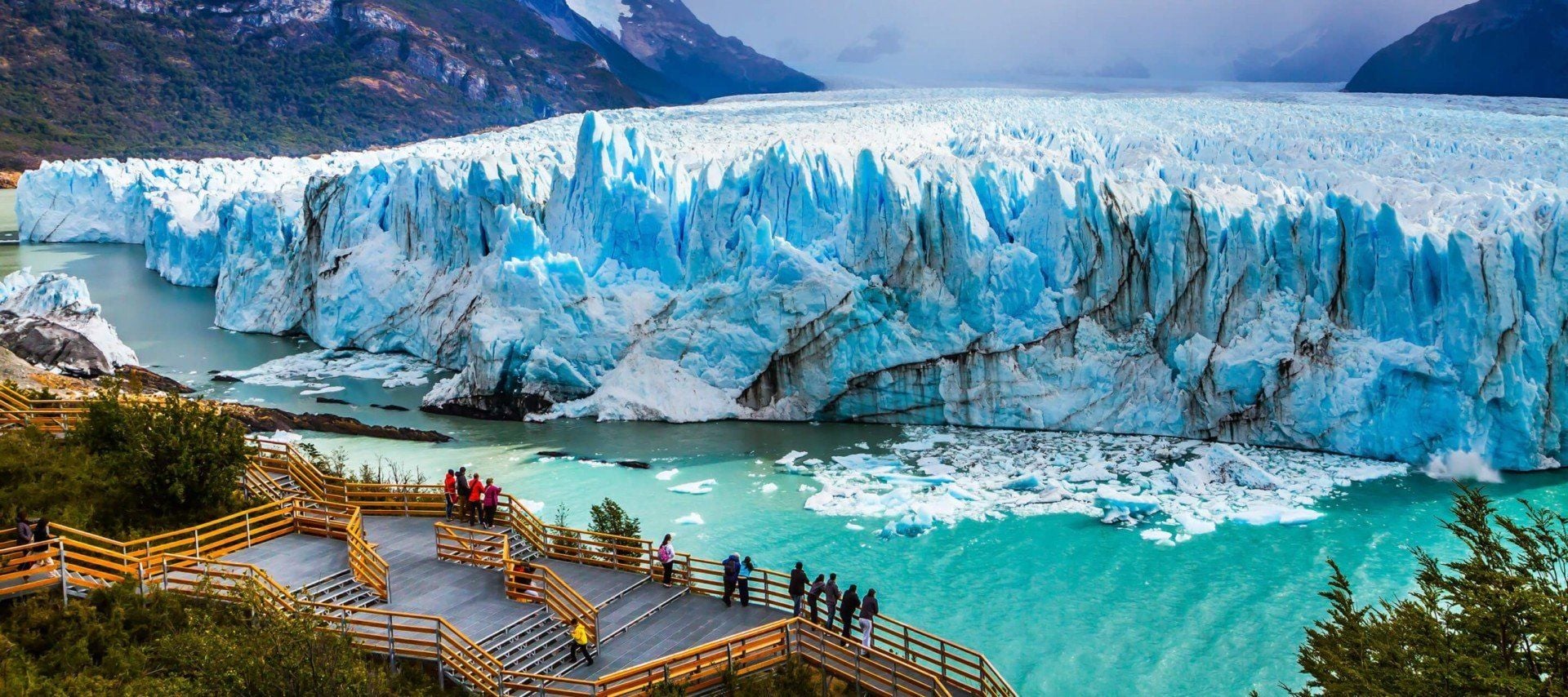 A group of people stand on a wooden observation deck overlooking a massive blue glacier meeting turquoise water in Patagonia, with mountains and greenery in the background—an unforgettable scene often highlighted on guided tours.