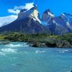 Turquoise river flows in the foreground with green vegetation on the banks, leading to dramatic, jagged Patagonia mountain peaks under a bright blue sky with scattered clouds.