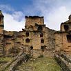 Ancient stone ruins with partially collapsed walls and towers invite you to experience a true adventure in Peru, surrounded by grassy paths and hills under a partly cloudy sky.