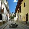 A narrow, cobblestone street ideal for South America travel, with historic buildings on both sides, a streetlamp, benches, and a small tree in the center. The sky is clear and blue with a few clouds above.