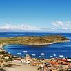 A scenic view of a coastal town with colorful buildings, boats floating on a blue bay, a small green peninsula, and distant mountains under a bright sky—perfect for capturing memorable moments.
