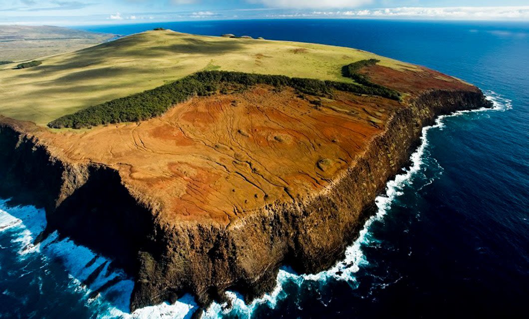 Aerial view of a coastal cliff on Easter Island with reddish-brown rocky terrain, bordered by green grass and trees on top, surrounded by blue ocean waves crashing against the base—an iconic scene ready for Explora adventures.