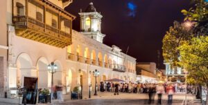 A lively night scene shows people gathering outside a historic colonial-style building with arches and balconies, warmly lit by street lamps, in one of the beautiful towns in Argentina’s bustling squares.