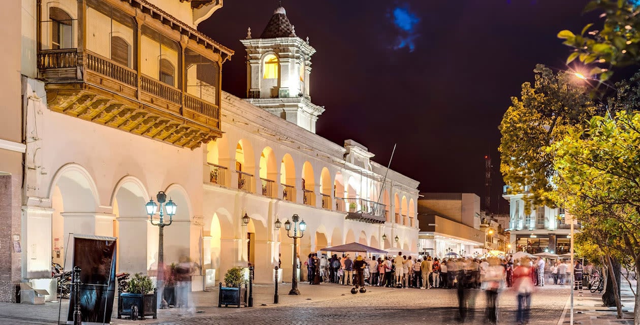 A lively night scene shows people gathering outside a historic colonial-style building with arches and balconies, warmly lit by street lamps, in one of the beautiful towns in Argentina’s bustling squares.