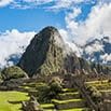Ancient stone ruins of Machu Picchu sit on green terraces with a steep, forested mountain and clouds in the background under a bright blue sky—a scene perfect for a fabulous trip.