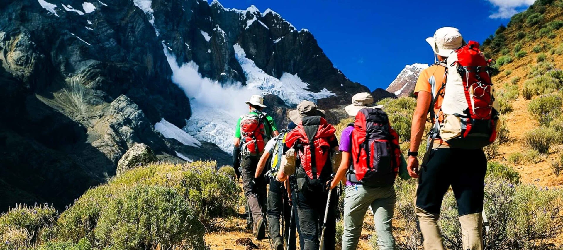A group of hikers with backpacks walk along a mountain trail toward snow-capped peaks, surrounded by rocky terrain and a clear blue sky—a perfect scene for Peru Adventure Tours.