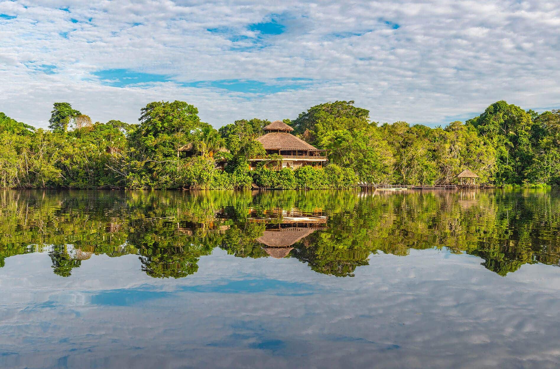 A wooden lodge with a thatched roof sits surrounded by lush green trees on the edge of a calm river, reflecting the building and forest in the water under a blue sky with scattered clouds.