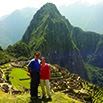 Two people stand smiling in front of the ancient Incan ruins of Machu Picchu in Peru, with green terraces and a steep mountain peak in the background under a bright sky—a perfect moment from their trip to Peru.