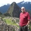 A man in a red shirt and sunglasses stands in front of the ancient ruins of Machu Picchu, where friendly guides ensure a well organized holiday amidst green terraces and the majestic mountain peak under a partly cloudy sky.