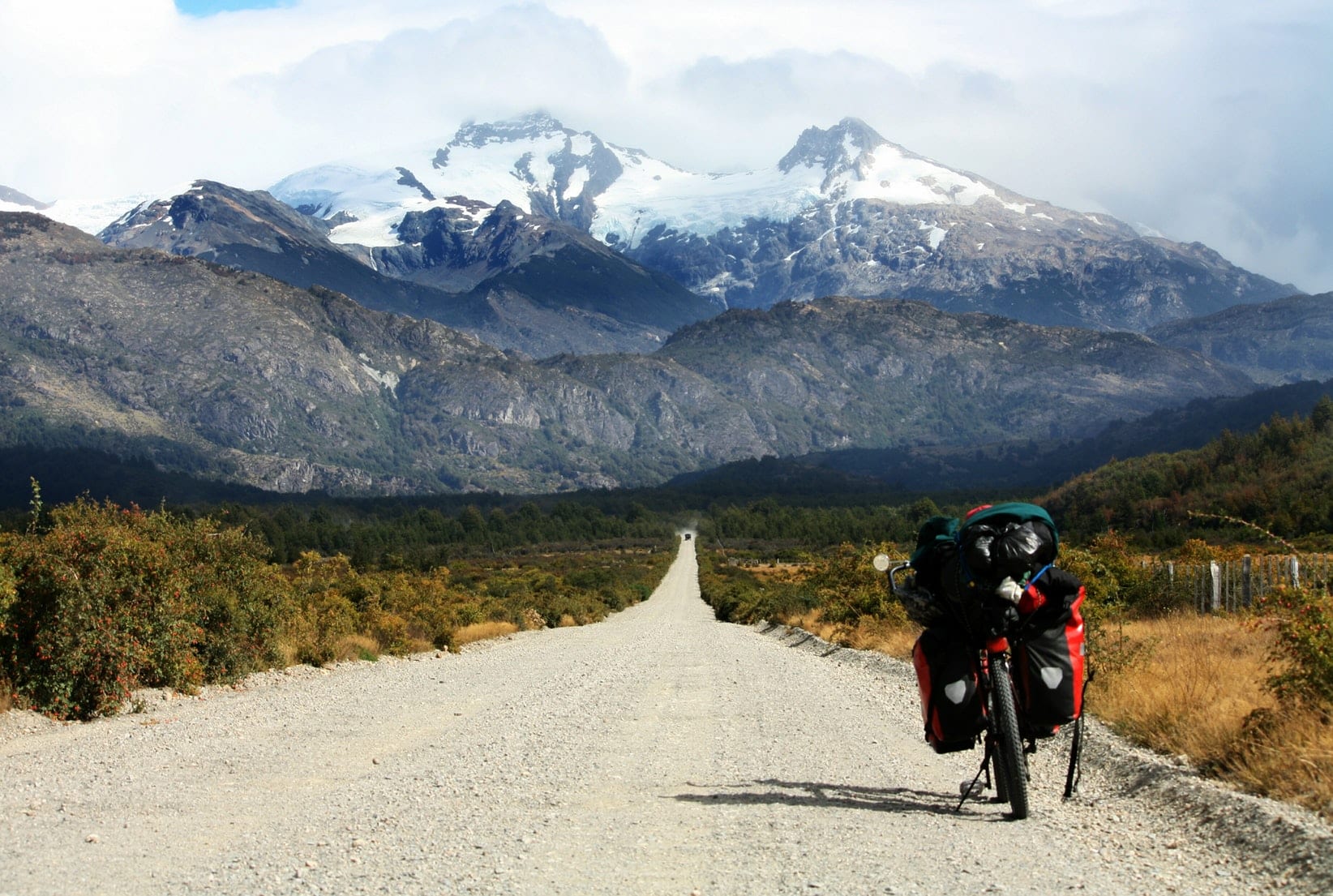 A loaded touring bicycle stands on a long gravel road leading toward distant snow-capped mountains, surrounded by greenery and rugged terrain under a cloudy sky.