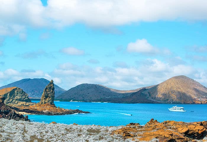 A scenic coastal landscape with rocky formations, blue water, and distant hills under a partly cloudy sky. A white boat sails on the calm sea near the shoreline—a perfect travel scene reminiscent of the Galapagos Islands.