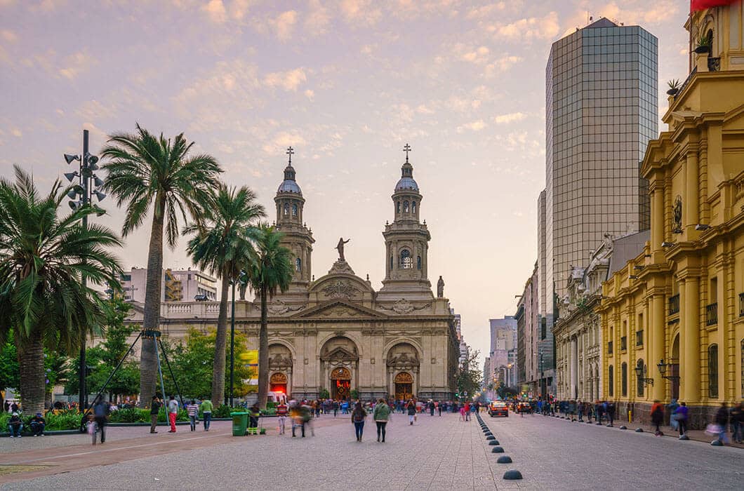 A bustling city square in Chile with palm trees, people walking, and a historic cathedral with twin towers in the center—surrounded by modern and classic buildings at sunset—a perfect spot to start exploring top Chile attractions.
