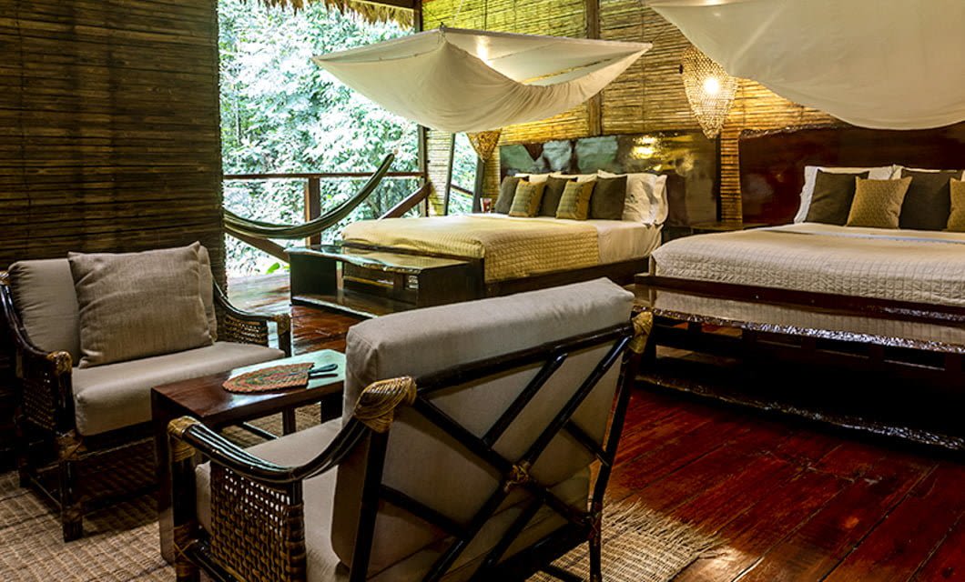 Cozy rustic bedroom at Posada Amazonas, a Peru Amazon Lodge, with two large beds, mosquito nets, cushioned chairs, a small table, and a hammock on a wooden veranda overlooking lush greenery. Natural wood textures create a warm, inviting atmosphere.
