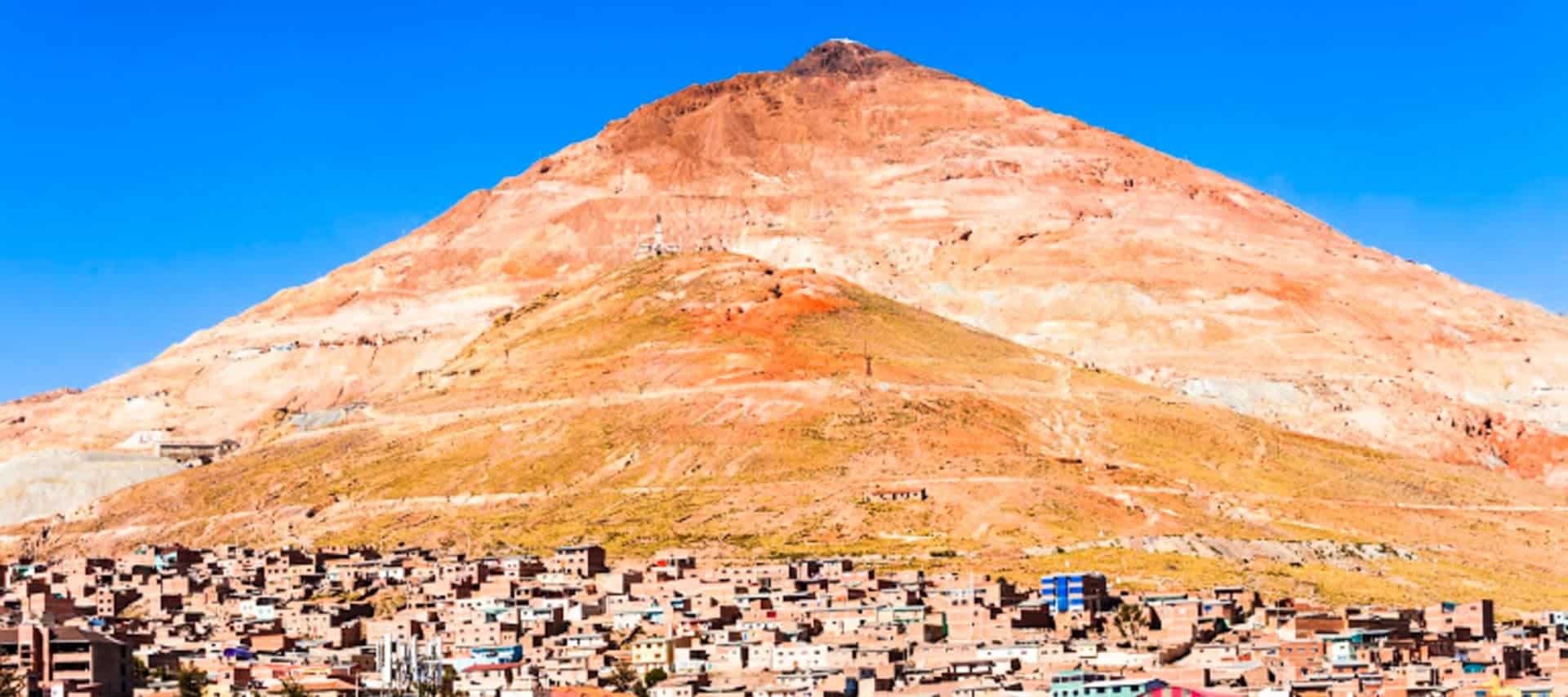 A large reddish-brown mountain with a rounded peak rises above a densely packed town of beige buildings under a clear blue sky, inviting travelers to discover its history with Potosi Tours.