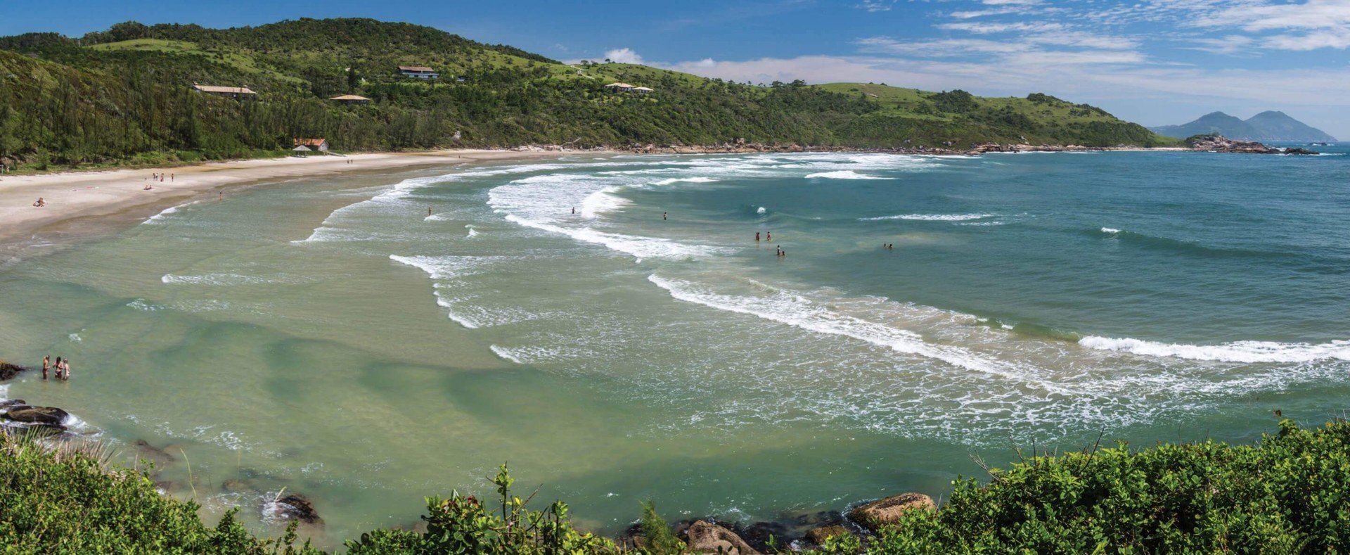 Wide sandy beach with gentle waves forming white lines, a few people in the shallow water, green hills with scattered houses in the background, and blue South America sky with some clouds hinting at pleasant weather.