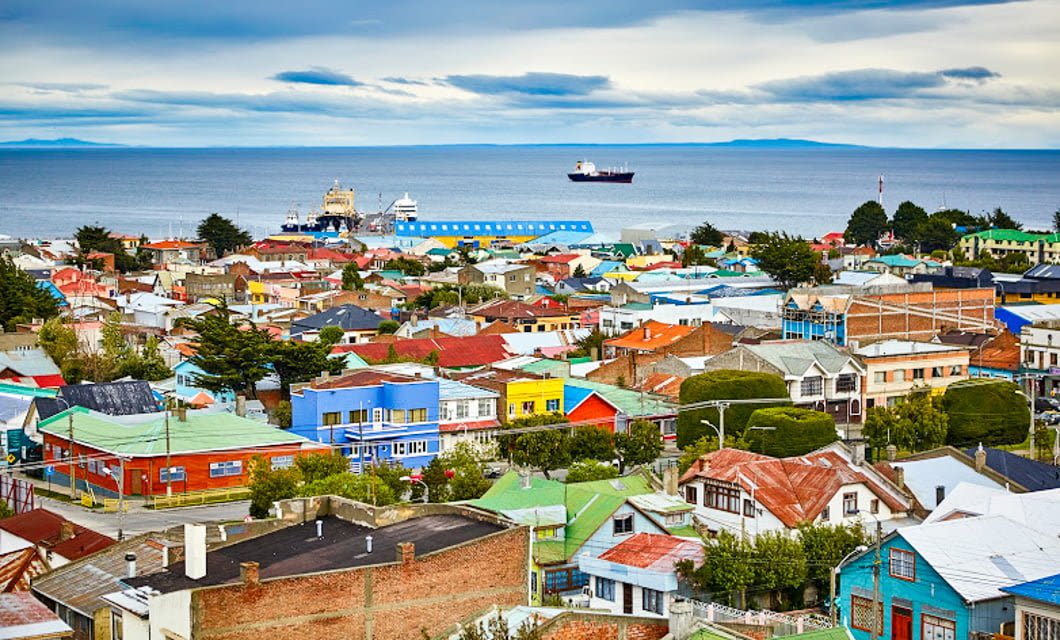 Colorful houses fill a coastal town with green trees; a ship and the Noi Indigo ferry are visible on the calm sea under a cloudy Patagonia sky.