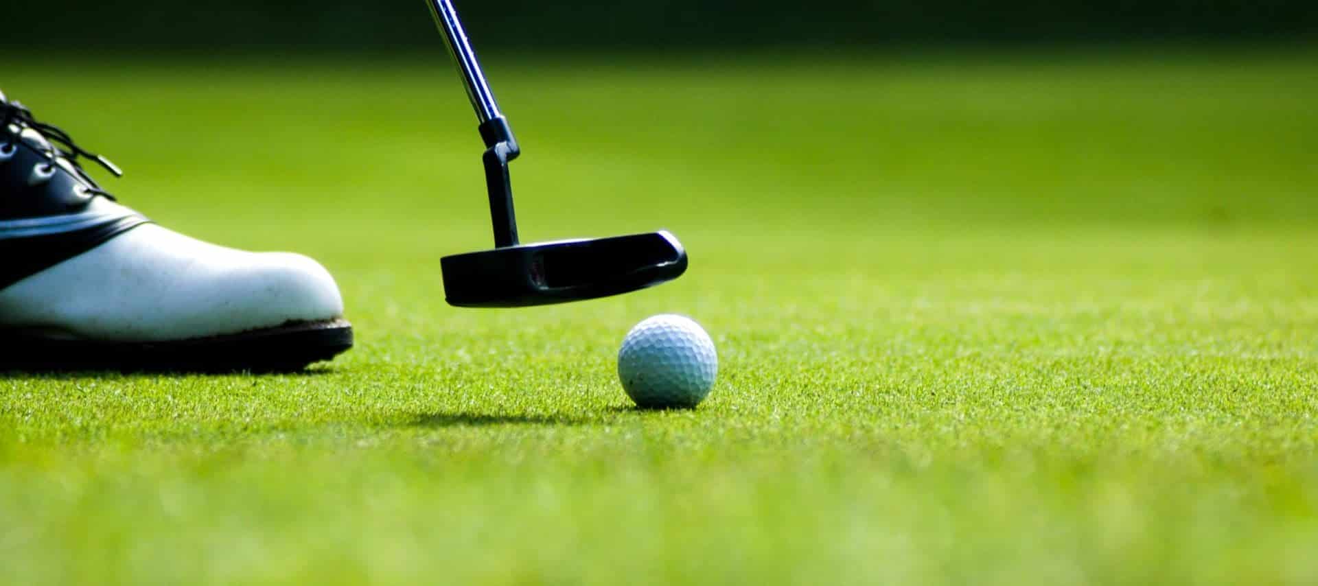 Close-up of a golfer’s shoe and putter about to strike a golf ball on a well-manicured green—perfect for Golf Tours in scenic regions like Patagonia. The focus is on the equipment, with a blurred grassy background enhancing the scene.