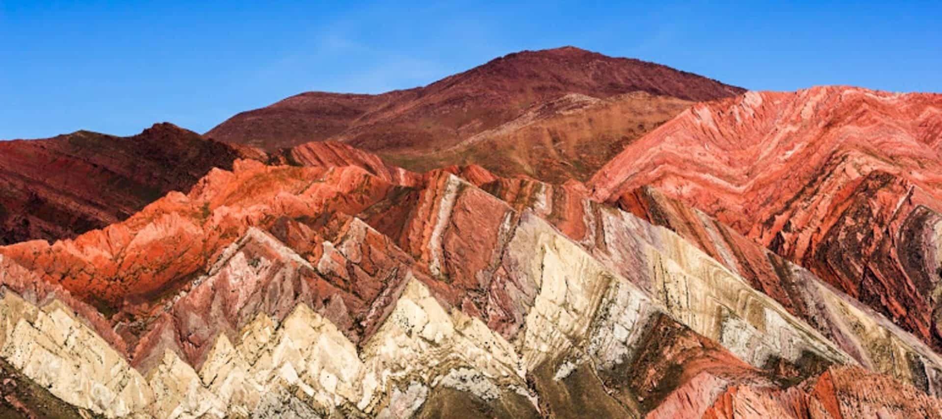 Colorful striped mountains with layers of red, pink, yellow, and white rock under a clear blue sky, featuring diagonal mineral bands in the rugged landscape of Quebrada de Humahuaca—perfect for unforgettable Quebrada tours.