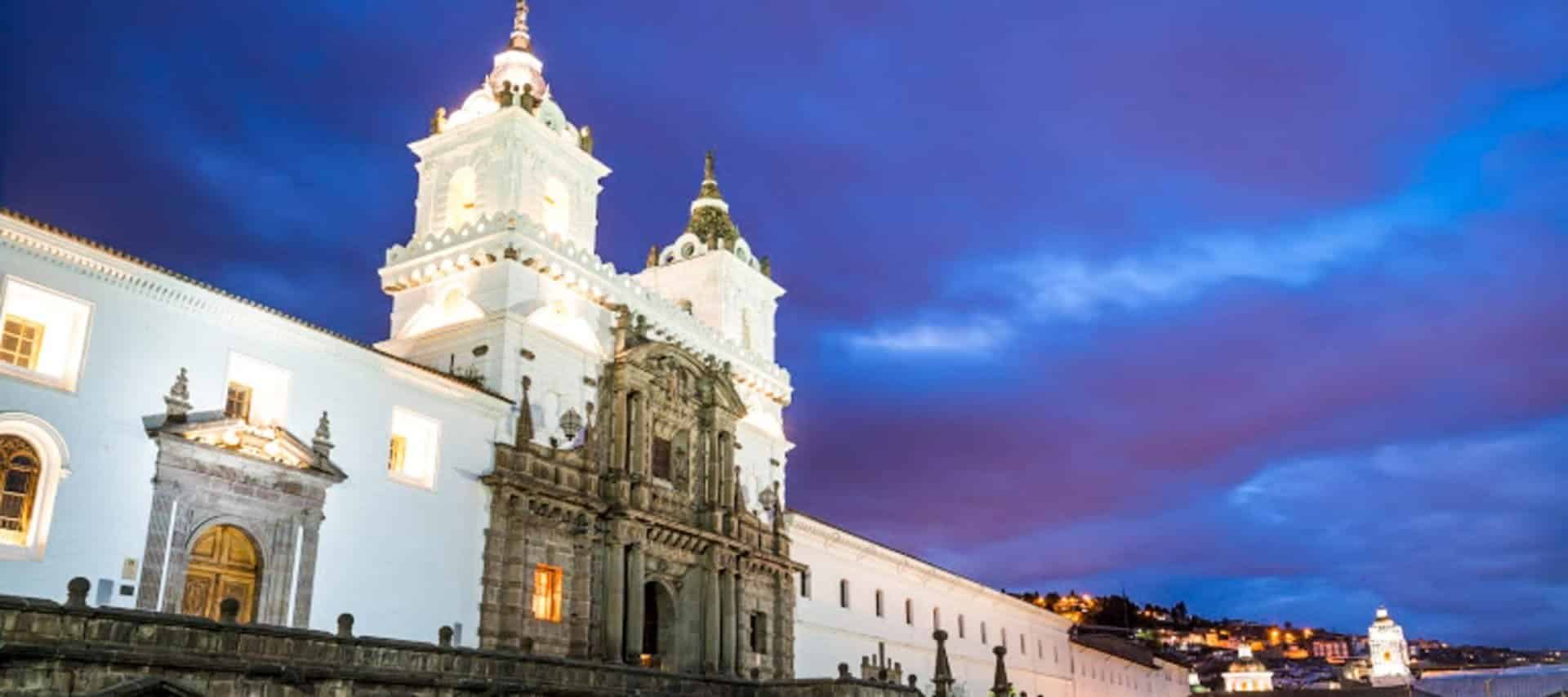 A historic white colonial church with two bell towers stands illuminated at dusk under a dramatic purple and blue sky in Quito, Ecuador—a must-see on Quito Tours of UNESCO heritage sites.